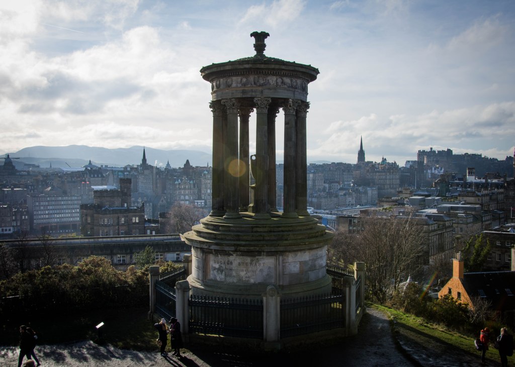 in the central foreground is a large pillared round structure on the plinth and surrounded by iron railings. A path curves around it which people are walking on. IN the distance are winter trees and the city of edinburgh with spires from churches and the castle. The sky is full of white cloud and there is lens flare in the centre of the image. 