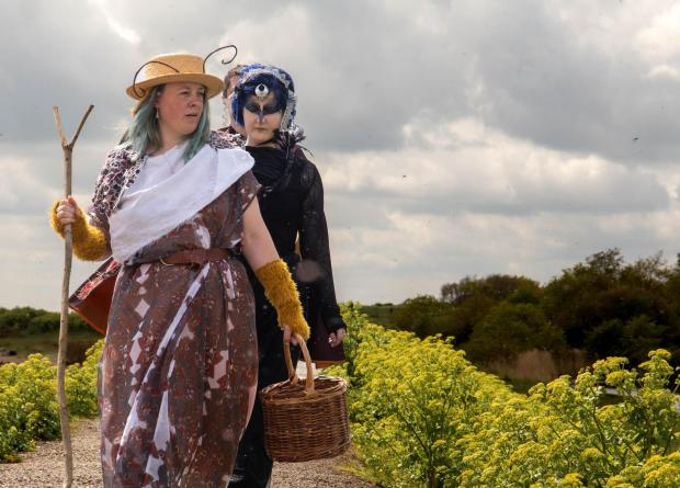 Two women walking along a country path surrounded by trees and shrubs. The woman in the front carries a staff and wears a hat. She has long pale turquoise hair and wears a brown robe. Behind her in black with a headress on and bird mask is a woman with dark blue hair. 