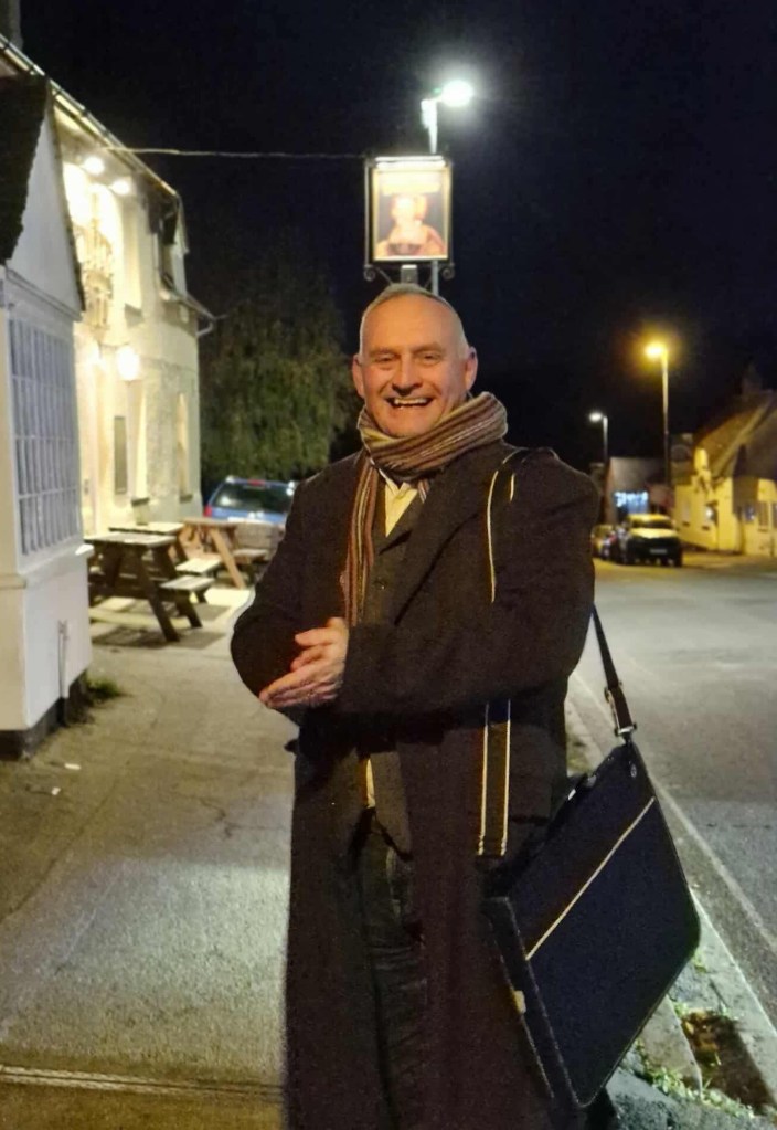 A man in a winter coat and scarf rubbing his hands for warmth he also wears a waistcoat and carries a large potfolio bag over his shoulder. He is smiling and the Queens Head pub is above his head. 