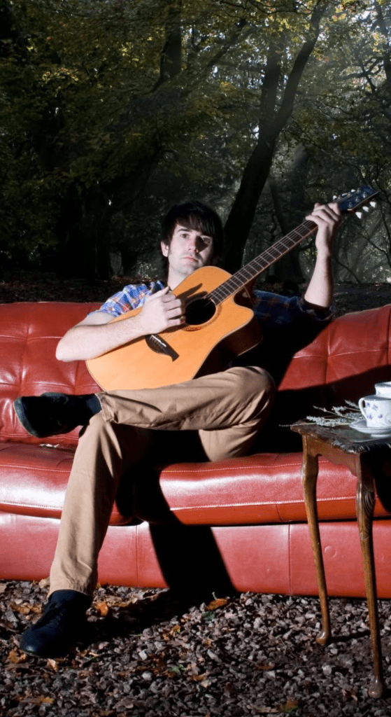 Olie campbell a young man playing a guitare whilst sitting on a settee in the woods. 