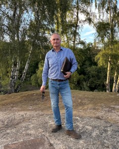 A man in a blue shirt and jeans staning below trees holding a portfolio under his arm.
