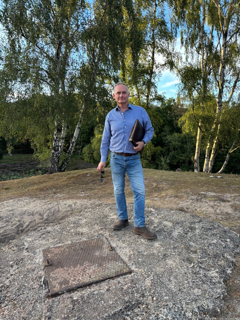 Bryan, wearing jeans and striped shirt, holding sun glasses and a portfolio under his arms is standing next to a metal hatch and is standing on poured concrete. Behind him are silver birch trees. 