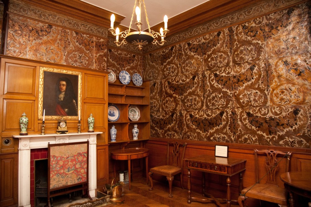 A wider image of the wallpapered room, from a distance the pattern on the wall paper resembles a grinning creature. The room has walnut brown futniture in it and a fireplace with a screen. There is a large painting of a 17th century man above the fire.