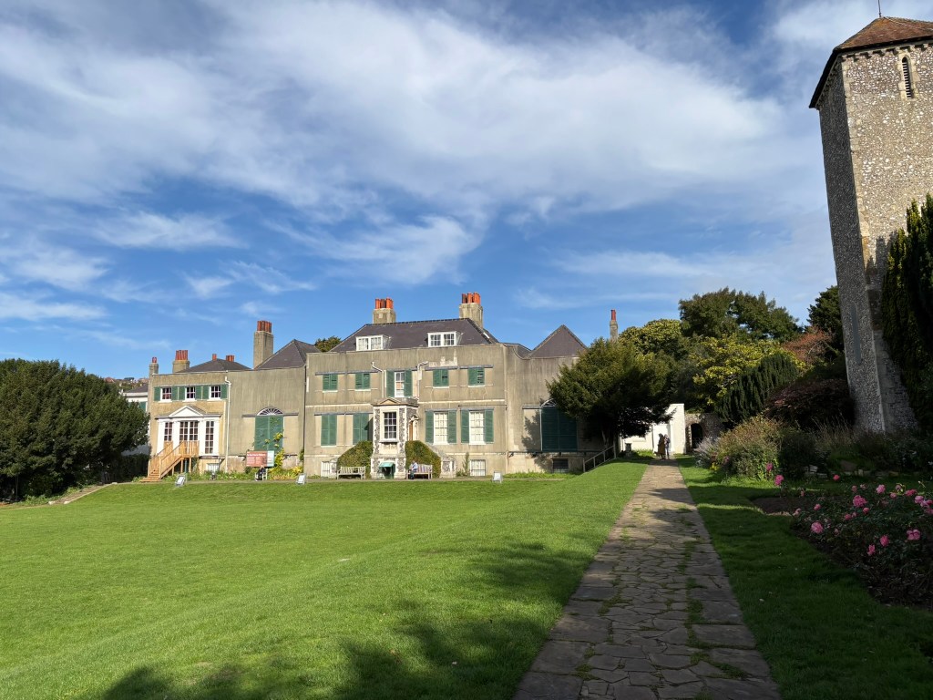 A pale grey house with green shutters and a wide flat green in front of it. The house has many chimneys and it's a bluesky sunny day.