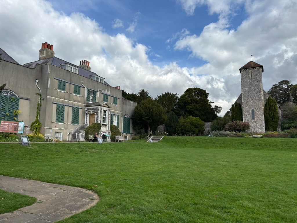 The back of the hosue grey with green shuttered windows and a croquet lawn in front to the right is a grey church tower