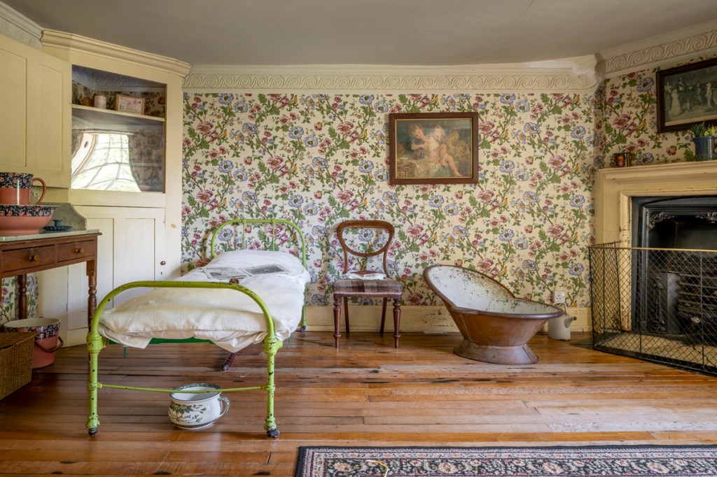 A flowery wallpapered bedroom with a simple metal bed and chamber pot below. A large fire and a hip bath in front.