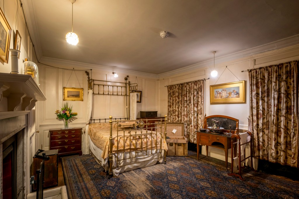A cream bedroom with flowery cutains and brass bedstead. A blue and gold patterend rug and walnut brown chest of draws and dressing table.