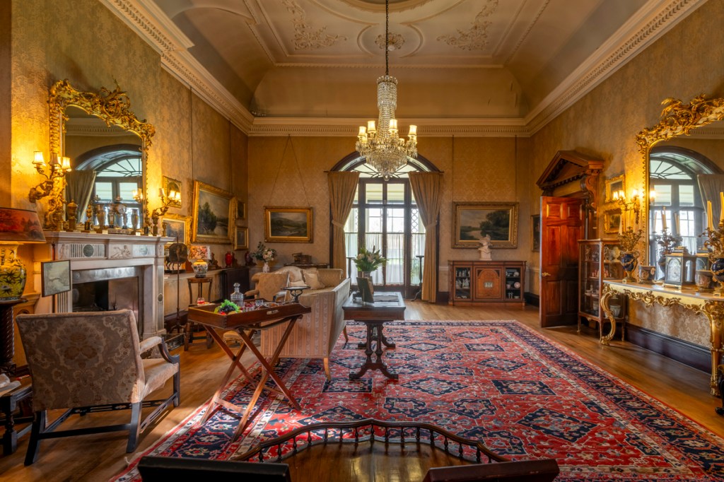 A room with gold framed mirrors, french doors at the end and a chandalier in the centre. Armchairs are by a fire and there is a large decoative rug on the floor.