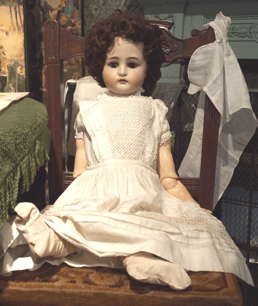 A large doll with china head and brown hair wearing a white dress sitting on a chair. 