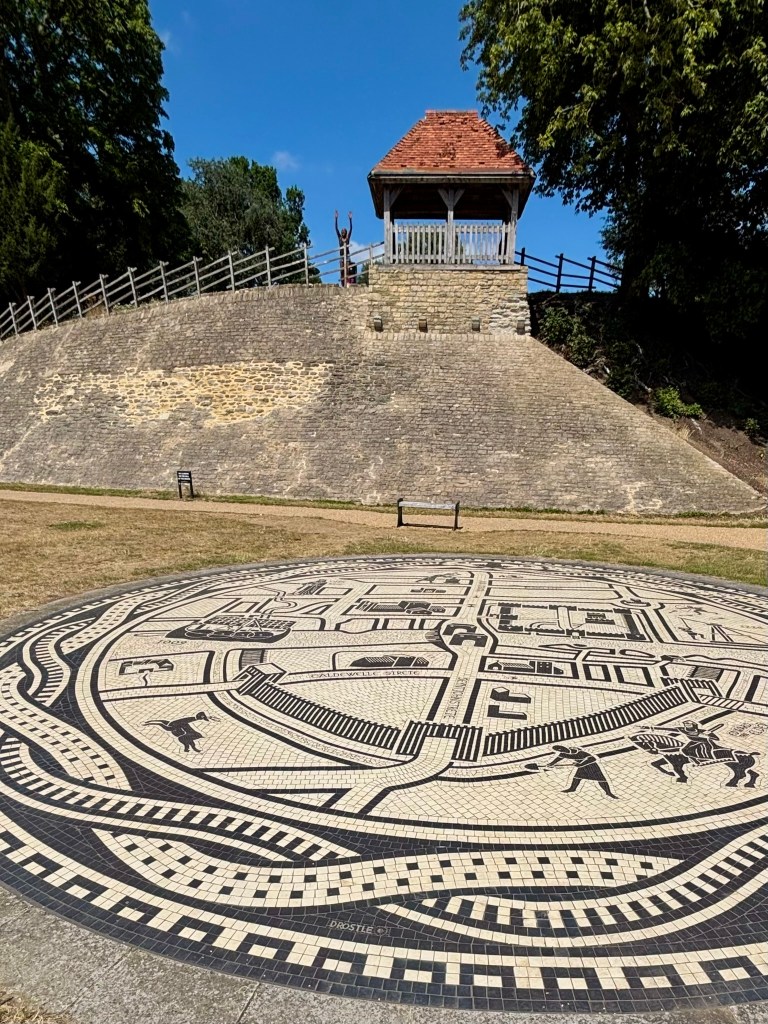 In the foreground a round mural of black and white on the ground showing people and animals like deer and horses. Behind it a stone mound with a railing going up it and small wooden porch at the top. Next to the porch a person is leaping into the air their hands above their heads. The mound is surrounded by trees. 