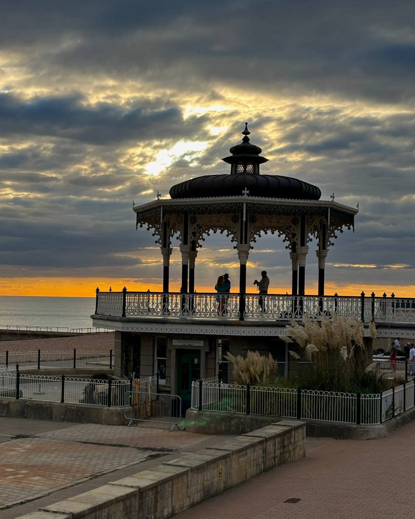 The Brighton seafront bandstand against the sea and sky at sunset. A deep orangey yellow strip of sky glows along the horizon and grey clouds are fringed with gold. ON the bandstand a couple are being filmed and are silhouetted against the sky. 