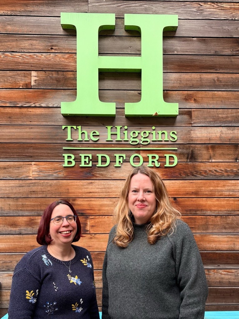 Two women standing beneath the Higgins sign on the left Lydia has red bobbed hair and on the right is Victoria with long blonde hair.