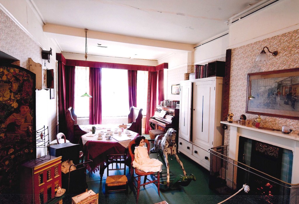 A small room with a green lino floor with burgundy curtains at the window. There's a round table set out for tea. A piano, a rocking horse, a dolls house and in the foreground there is a chair with a doll with dark hair sitting on it. 