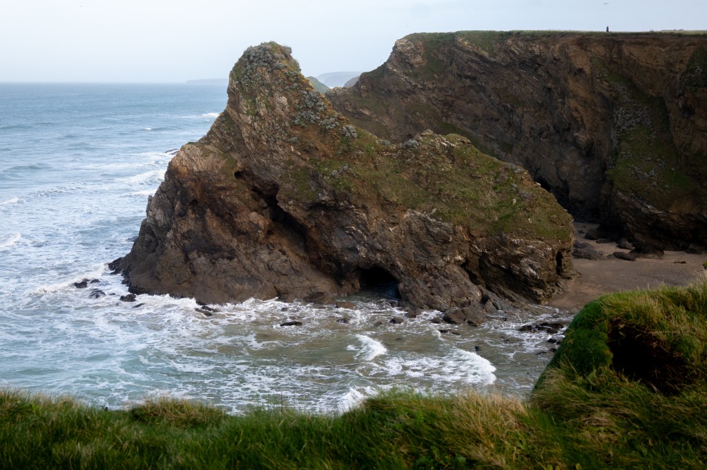 In the centre Black Humphrey's rock which is jagged and has a rounded point on top. There are caves underneath the rock and along it's sides. Behind it to the right is a large cliff. To the left is sea and the sky is pale blue.