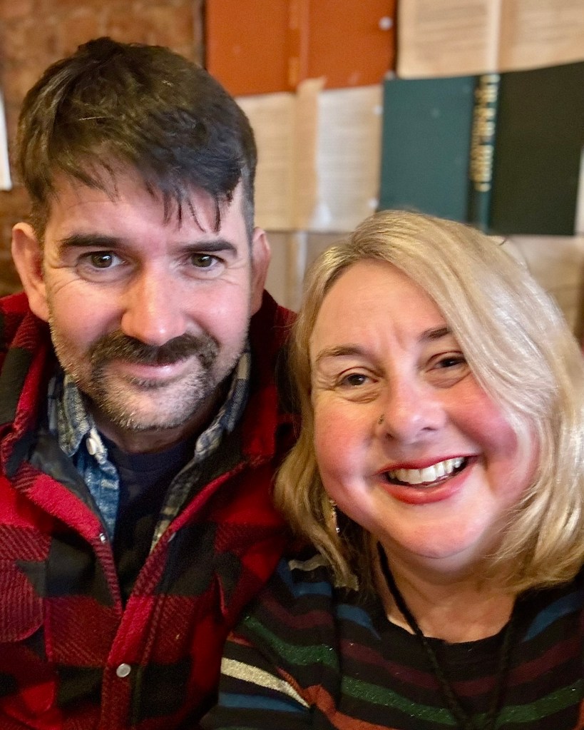 Olie a man with dark hair and eyes and a moustache and beard wearing a red checked shirt and me a woman with blonde hair smiling. I'm wearing a stripy jumper. 