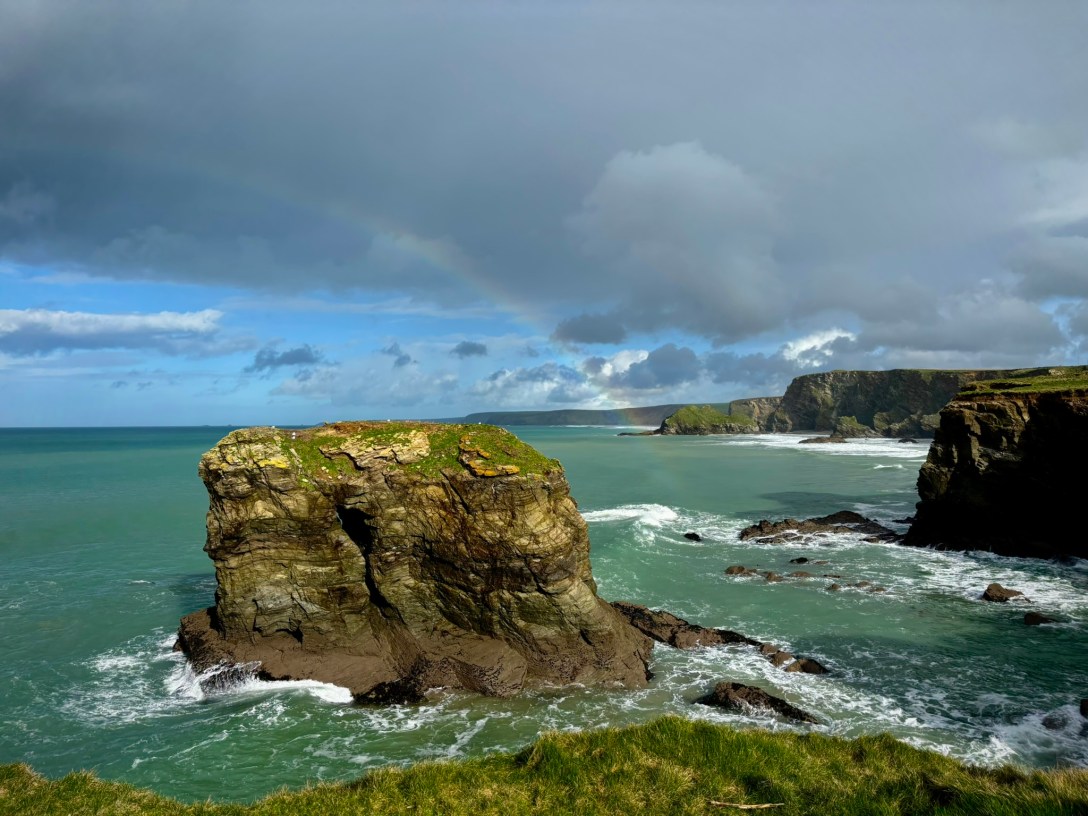 A deep turquoise coloured sea with jagged cliffs to the right and in the centre a small rocky island. The sky is blue with grey clouds.