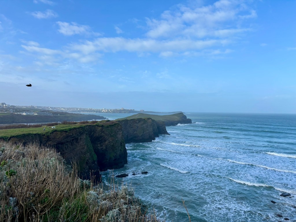 The edge of whipsiderry cove with waves rushing in. IN the middle distance is the undulating headland of Trevelgue Head on Porth Island. In the distance the sun is shining on Newquay. The sky and sea are a pale baby blue colour.
