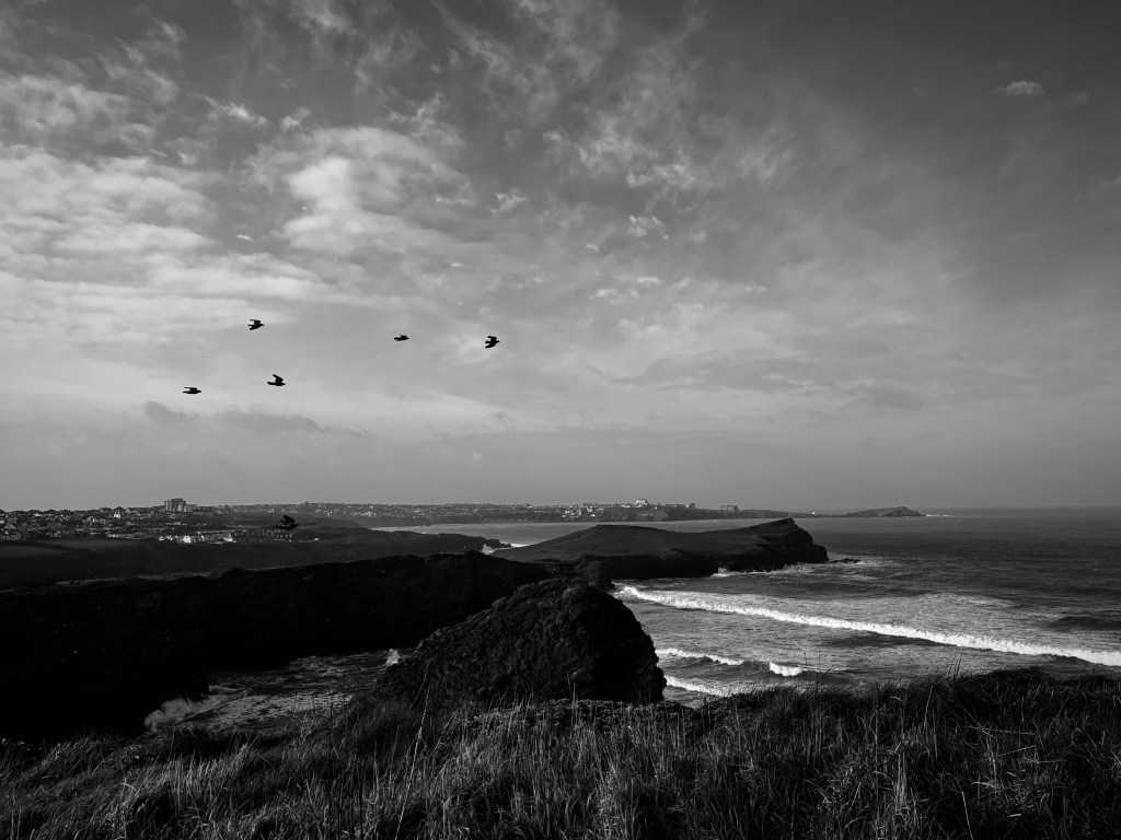 A black and white image of Whipsiderry cove, with waves rushing in. The top of Black Humphrey's rock can be seen above the headland in the foreground. IN the middle distance is Porth Island / Trevelgue head and then in the far distance the headland at Newquay. IN the sky are jackdaws flying.