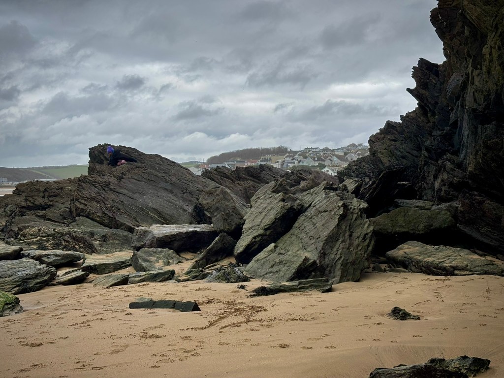 A collection of jagged and sharp rocks next to a cliff. There is sand in front and bits of seaweed. IN the distance are houses and a grey sky. There is a child wearing a purple hat on one of the rocks.