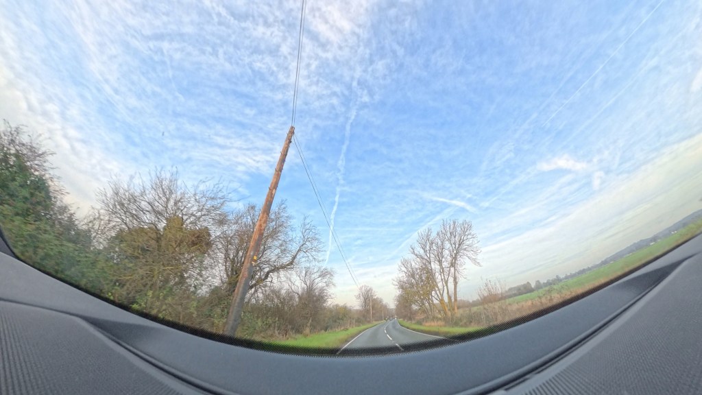 Dashcam image of road gently curving with a telegraph pole and some trees.