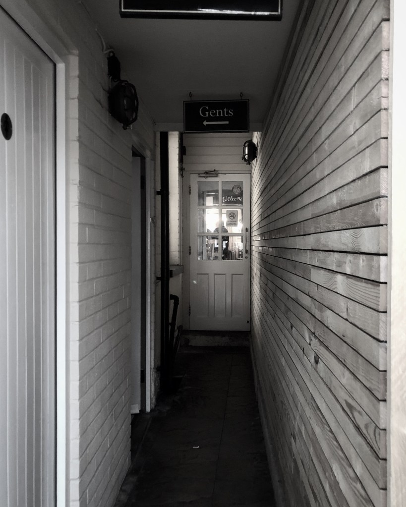 A narrow passage with stone flags as the floor and the sign to the gents toilet. 