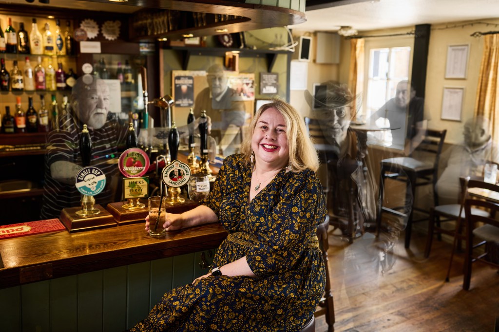 Nat sitting on a bar stool at the bar smiling she is surrounded by semi translucent mainly older men.