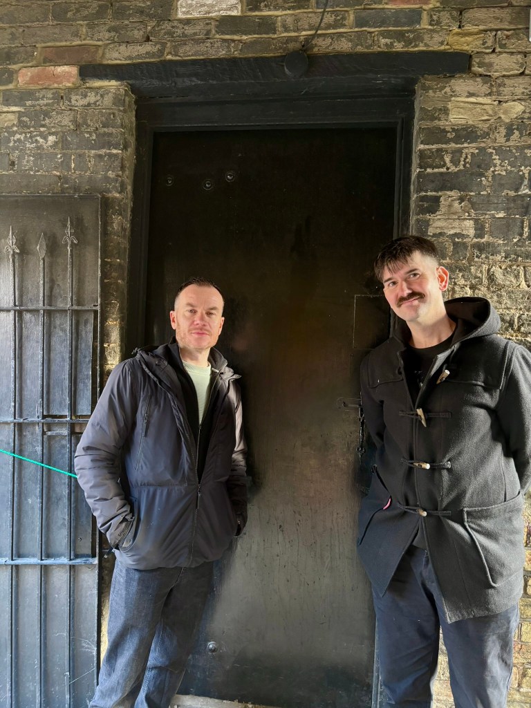 Two men Mikey and Olie standing either side of an old black door in a brick shed.