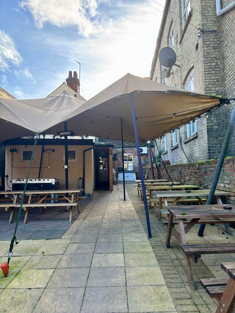 The whole of the back yard of the pyb with a canopy above some of it and rows of picnic style tables and a subuteo machine. 