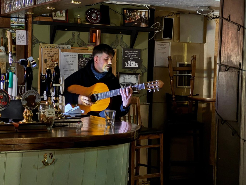 Olie a tall man with dark hair and moustache playing acoustic guitar sitting at a wooden bar in a dark pub the only light is on him.