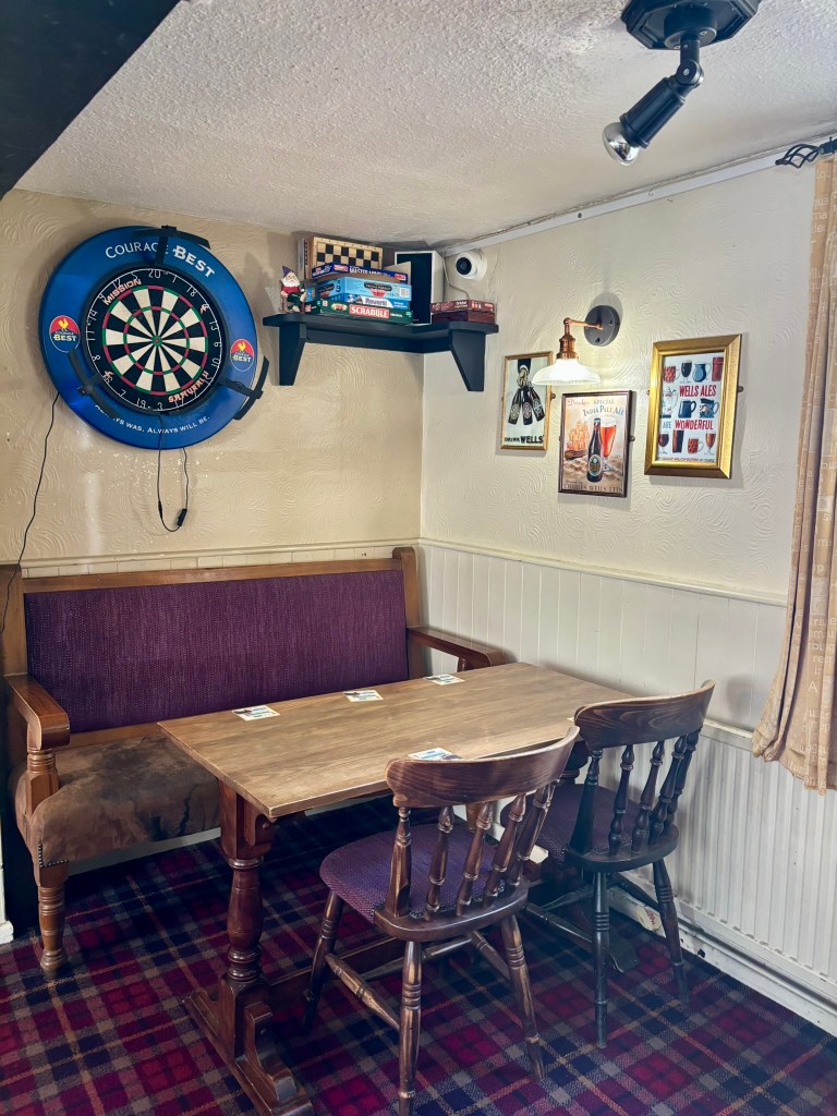 A settee  with rectangular table in front and chairs. Above the settee is a dart board and shelf. (There is a gnome in a rocking chair on the shelf.)