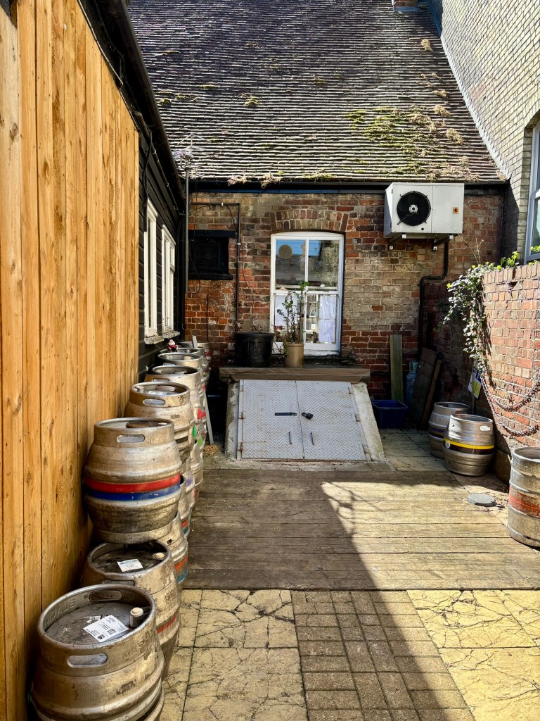 The back of the pub looking towards the cellar doors which are in the ground at a slope. There are empty barrels around and a window into the pub. 