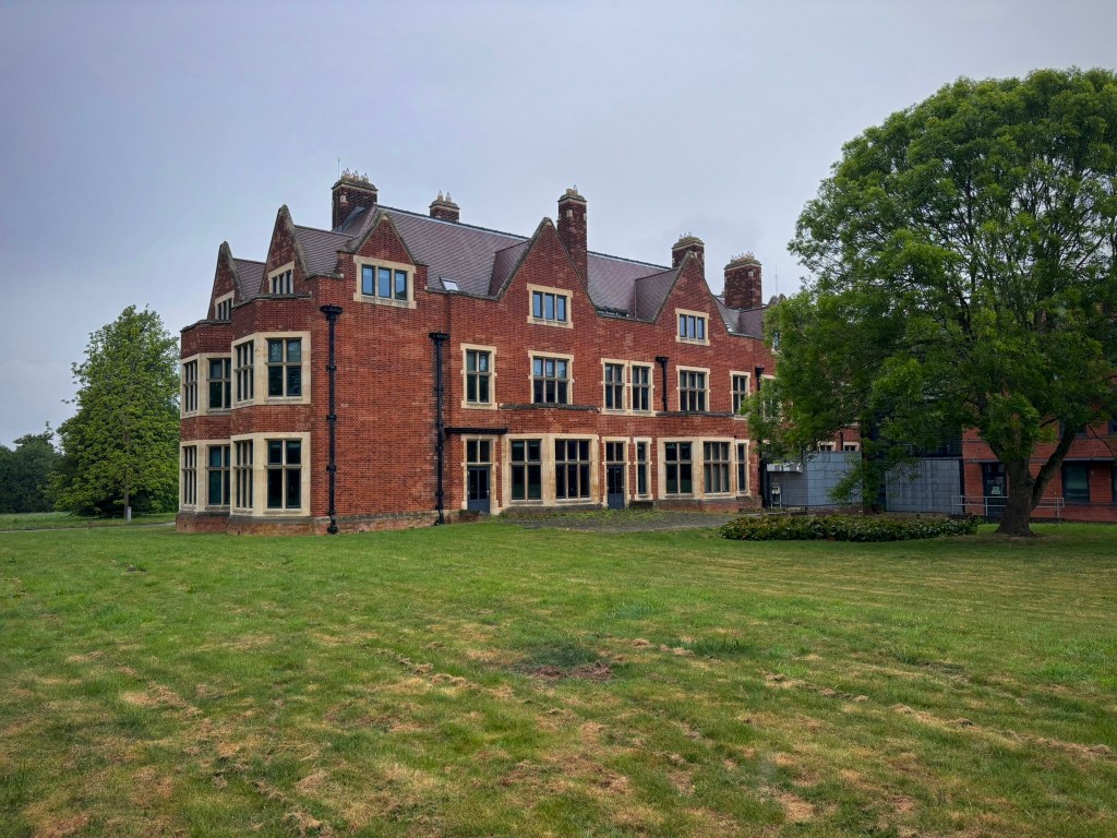 A large red brick manor house with lawn and side extension also in red brick but more modern. Large trees surround the manor. 