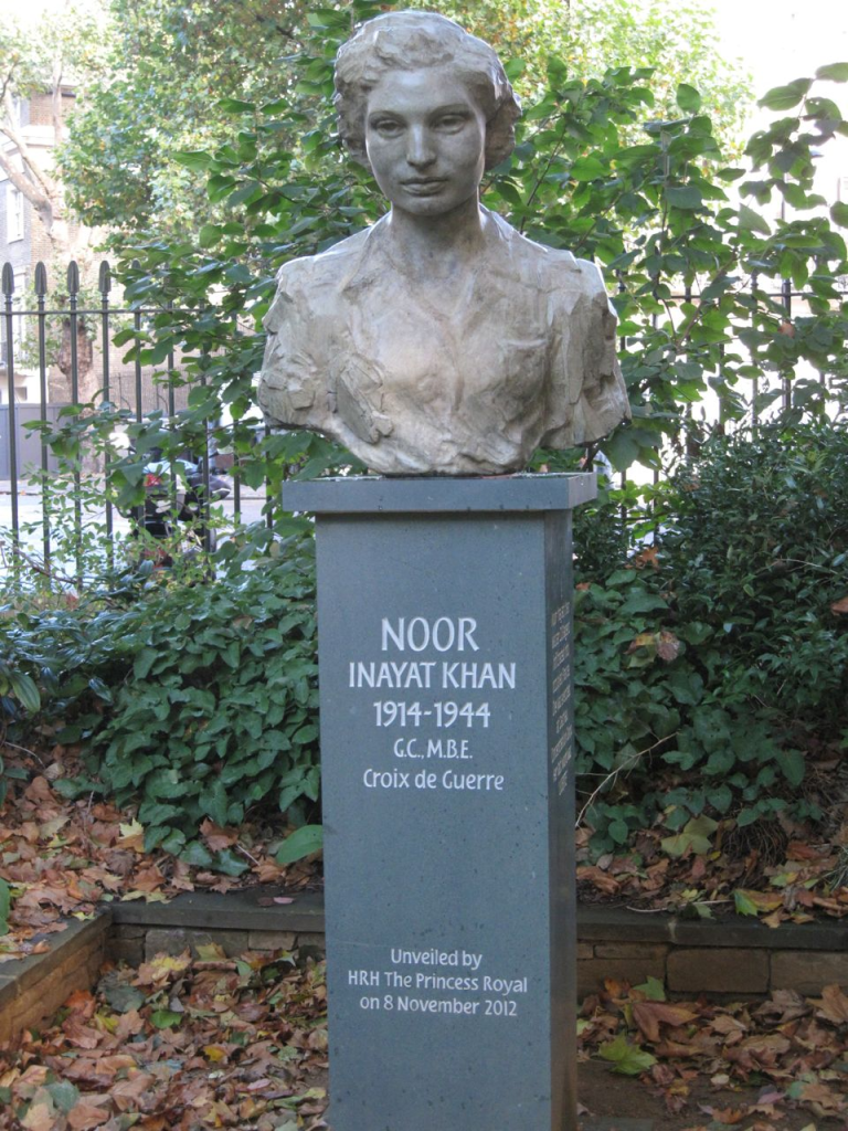 A bronze bust of a young woman with wide mouth and kind expression on a plinth with a backdrop of iron railings and plants. 