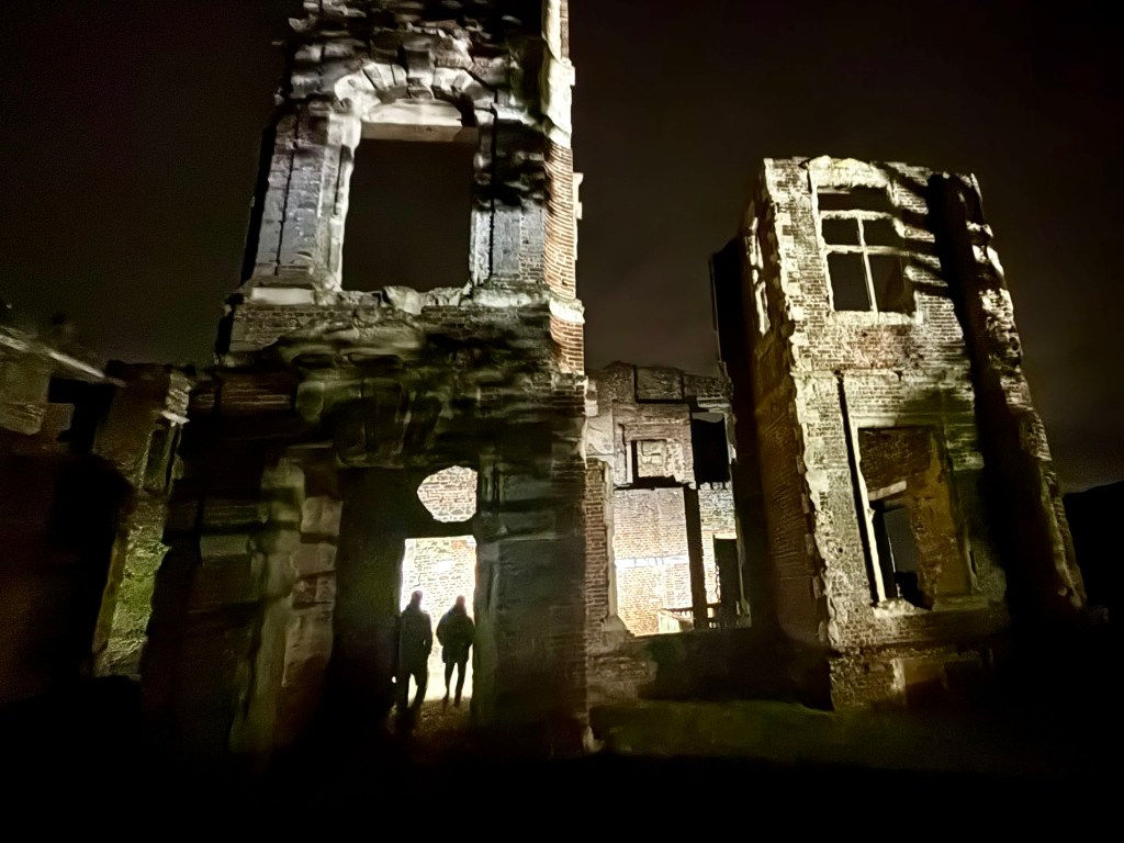 The gothic façade of a ruined house at night with large gaping windows and doors and ragged towers with no roof. Two shadowy figures are entering the house lit up by their torch light. 