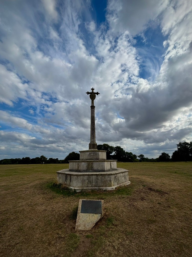 A large stone gothic cross on an octagonal plinth with plaque in front. Behind are trees and grey clouds with patches of blue sky