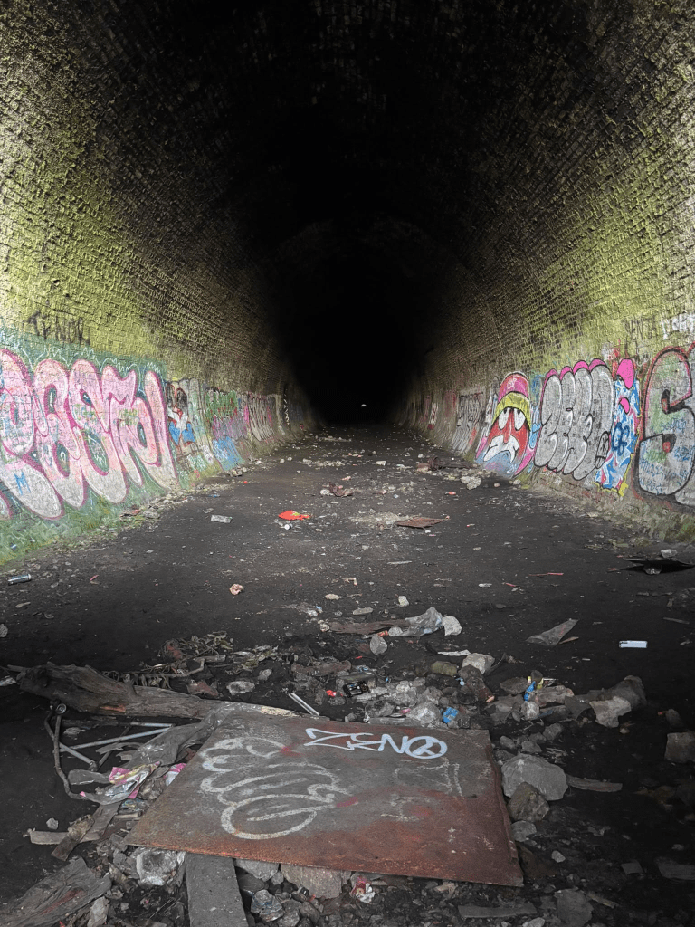 A dark, arched brick tunnel with walls covered in colorful graffiti tags in pink, blue, and white. The floor is littered with debris, rubble, and a rusted metal sheet with graffiti, and the passage fades into darkness with a faint point of light visible far in the distance.