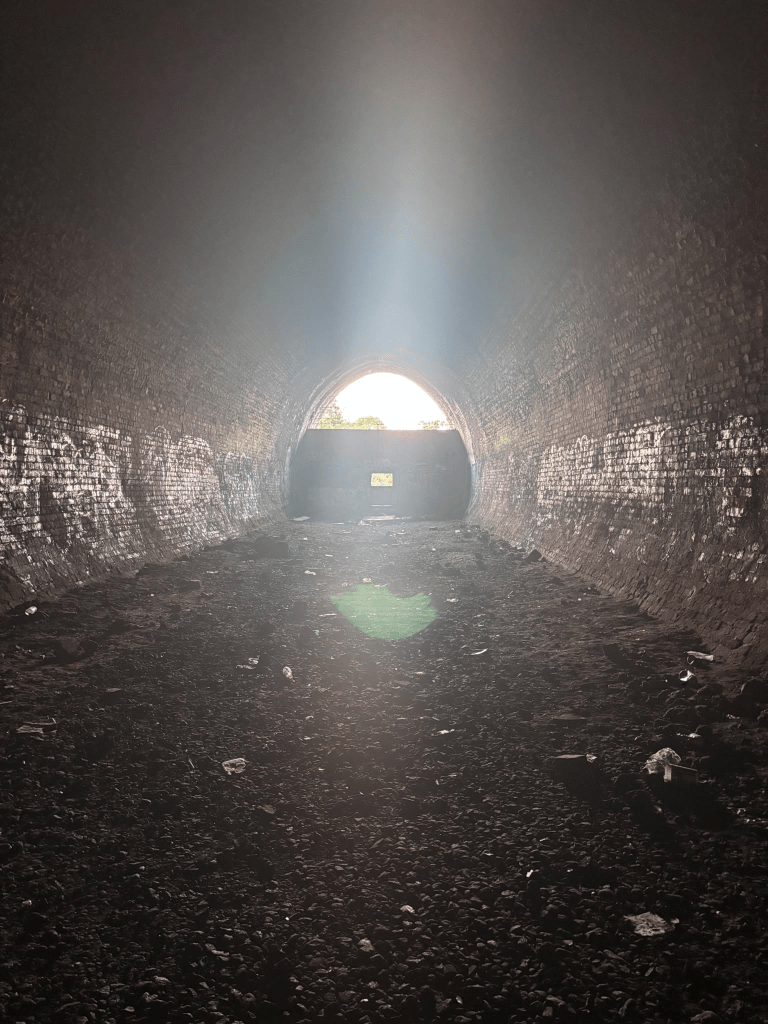 The dark interior of a graffiti-covered brick tunnel, with a shaft of bright light streaming in through the arched opening at the far end. A wooden barrier with a small square gap partially blocks the exit, and litter and debris cover the tunnel floor.