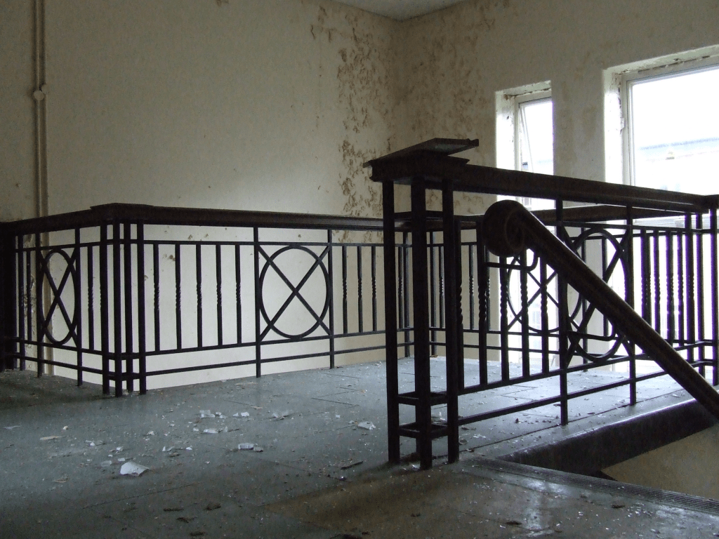 An art deco style staircase of wooden banister and metal posts, The floor is grey and covered with debris and flakes of paint.