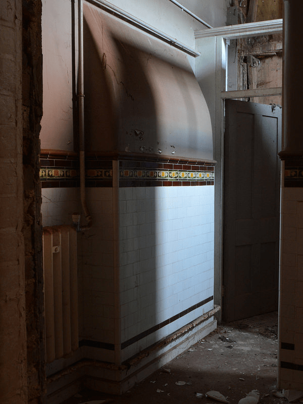 The interior of a derelict building showing a wall lined with white tiles and a decorative patterned border, with an old cast-iron radiator below. Peeling paint and exposed brick are visible on the surrounding walls, and light streams in through an open doorway leading to another room