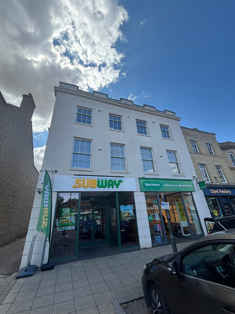 A three-storey white-painted building on a high street, with a Subway sandwich shop and a Specsavers store at ground level, and a Card Factory shop visible to the right. Sash windows line the upper floors, and a car is parked on the street in front under a partly cloudy blue sky