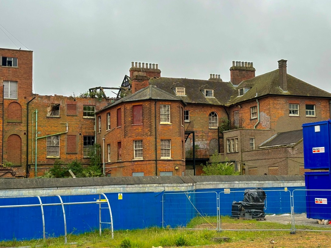 A derelict red-brick Victorian building under a grey overcast sky, its left section gutted by fire with collapsed roof timbers, empty window frames, and exposed brickwork, while the right section remains more intact with rows of boarded and broken windows and multiple chimney stacks. A blue construction hoarding with yellow warning signs and a metal fence run along the front of an overgrown grassy bank.
