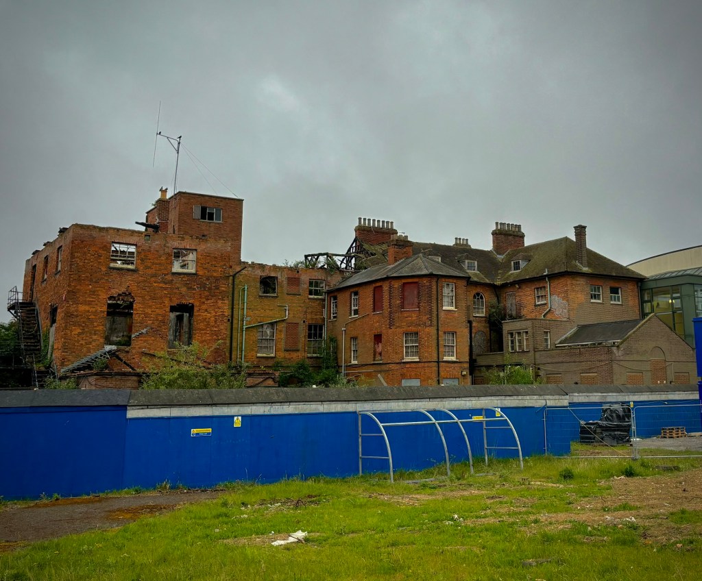A derelict red-brick Victorian building under a grey overcast sky, its left section gutted by fire with collapsed roof timbers, empty window frames, and exposed brickwork, while the right section remains more intact with rows of boarded and broken windows and multiple chimney stacks. A blue construction hoarding with yellow warning signs and a metal fence run along the front of an overgrown grassy bank. 
