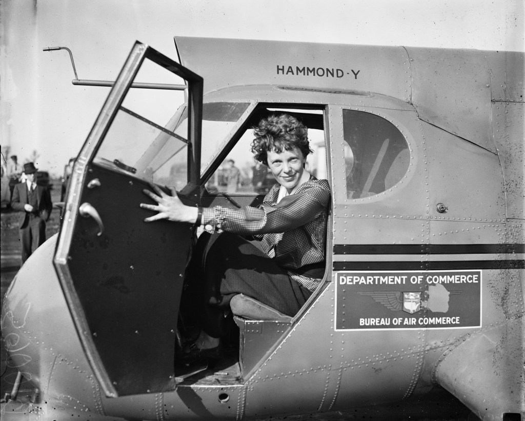 Amelia Earhart sitting in a plane with the door open. The plane has department of commerce written on its side. Amelia is wearing a checked shirt and is smiling. 