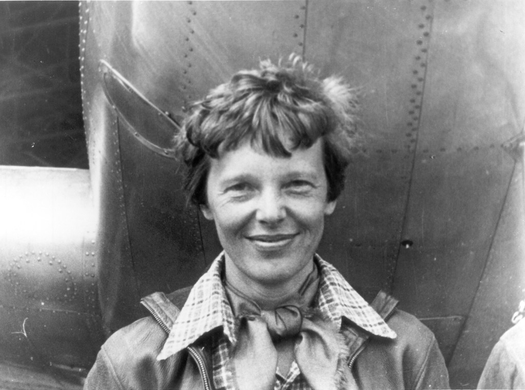 Amelia Earhart, smiling underneath the nose of her plane. She has short tousled hair, a close lipped smile and is wearing a scarf tied at her neck, a plaid short and flying jacket. 