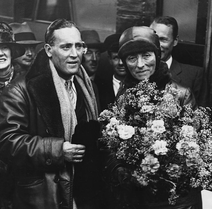 a man in a flying jacket and scarf with slicked back hair on the left on the right an older woman with a flying cap holds a huge bunch of flowers she is smiling.