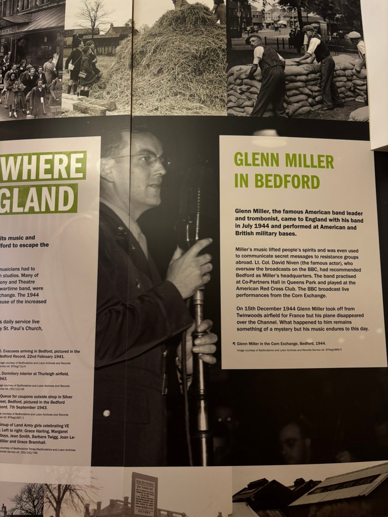 An information board with an image of Glenn Miller wearing glasses and holding his trombone. The sign next to it reads Glenn MIller in Bedford, 