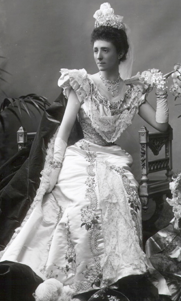 The Duchess in evening gown with a hat of flowers on her dark hair, the dress is very fancy with a sinched in waist, flouncy shoulders but sleeveless. She has long evening gloves with embroiders flowers as does her dress skirt. 