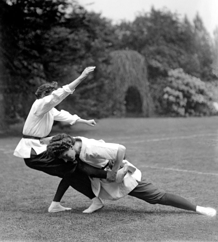 Two women competing in jujitsu the woman on the right is lunging forward and  has hold of the woman on the left's leg, The woman on the left has her arms in the air and is falling backwards. Both women have hair plaited and wrapped around their heads and are wearing dark loose trousers and white shirts with belts. 