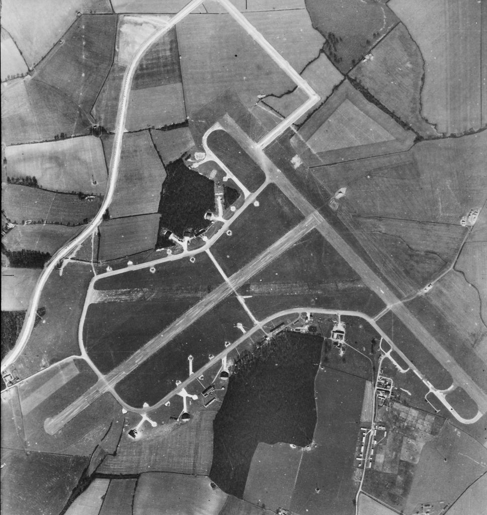 A black and white aerial photograph of fields and runways, huts and small buildings are scattered about the edge of the runways. 