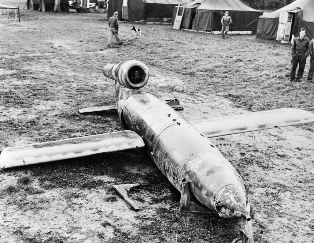 A Doodle bug rocket with two wings, tale, pointed front and raised section above the tail. It is in a field there are men in the background and a black and white dog.